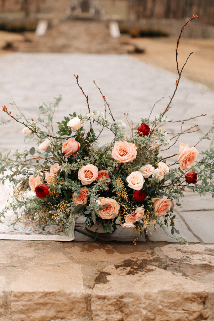 Organic growing wedding ceremony urns in floral hues of dusty pink, red, pale yellow, and sage green composed of petal heavy roses, garden roses, eucalyptus, winter branches, and natural greenery. Design by Rosemary and Finch in Nashville, TN. 