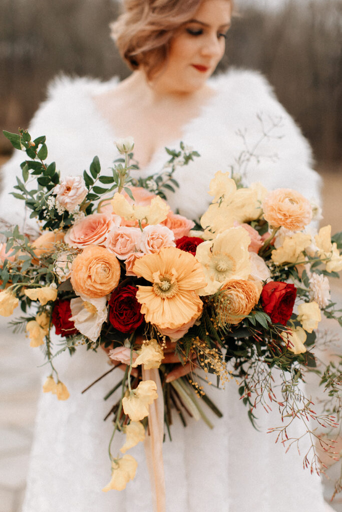 Elegant winter wedding bridal bouquet composed of Icelandic poppies, garden roses, ranunculus, butterfly ranunculus, sweet peas, jasmine, spray roses, and natural greenery in floral hues of peach, pale yellow, red, dusty pink, and sage green. Design by Rosemary and Finch in Nashville, TN.