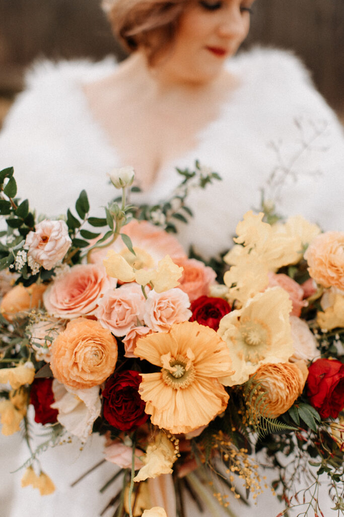 Elegant winter wedding bridal bouquet composed of Icelandic poppies, garden roses, ranunculus, butterfly ranunculus, sweet peas, jasmine, spray roses, and natural greenery in floral hues of peach, pale yellow, red, dusty pink, and sage green. Design by Rosemary and Finch in Nashville, TN.