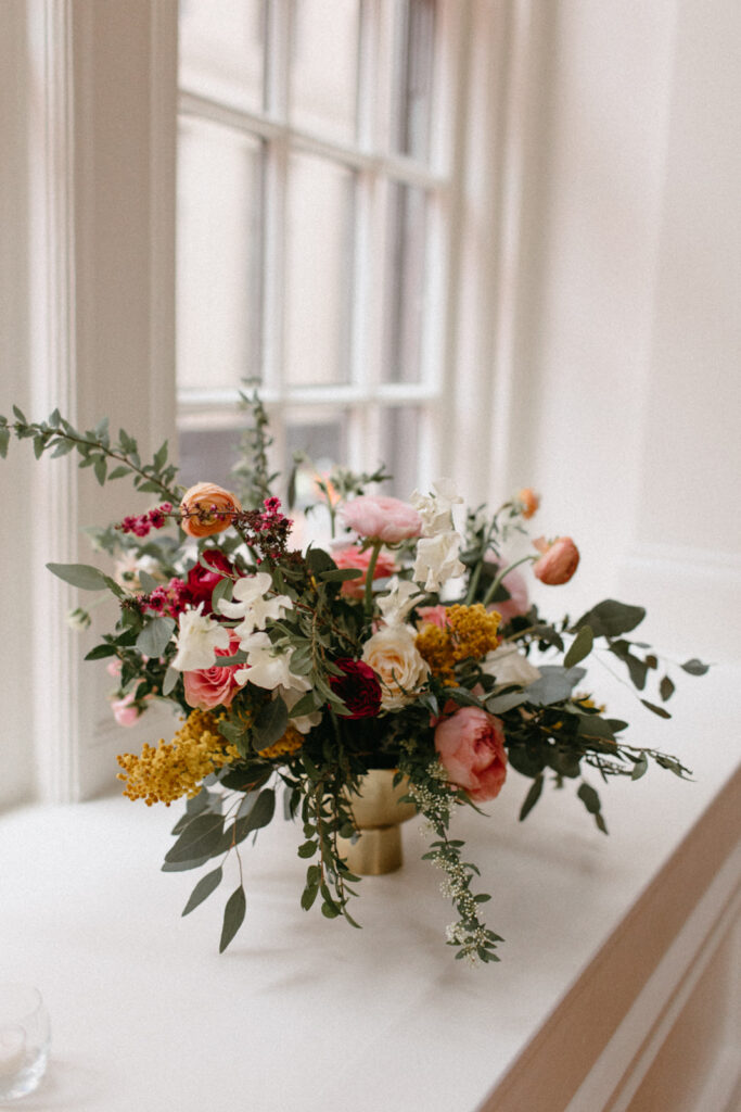 Elegant window floral arrangement ascent the winter ceremony space in hues of burgundy, mustard yellow, white, pink, and sage green composed of garden roses, sweet peas, acacia, ranunculus, spirea, and eucalyptus. Design by Rosemary and Finch in Nashville, TN. 