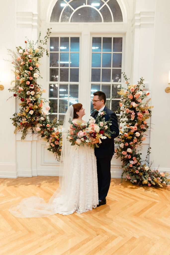 Asymmetrical growing floral installation for winter wedding in hues of mustard yellow, pink, burgundy, white, and sage green composed of petal heavy roses, acacia, mums, spray roses, butterfly ranunculus, ranunculus, and natural greenery. Design by Rosemary and Finch in Nashville, TN. 
