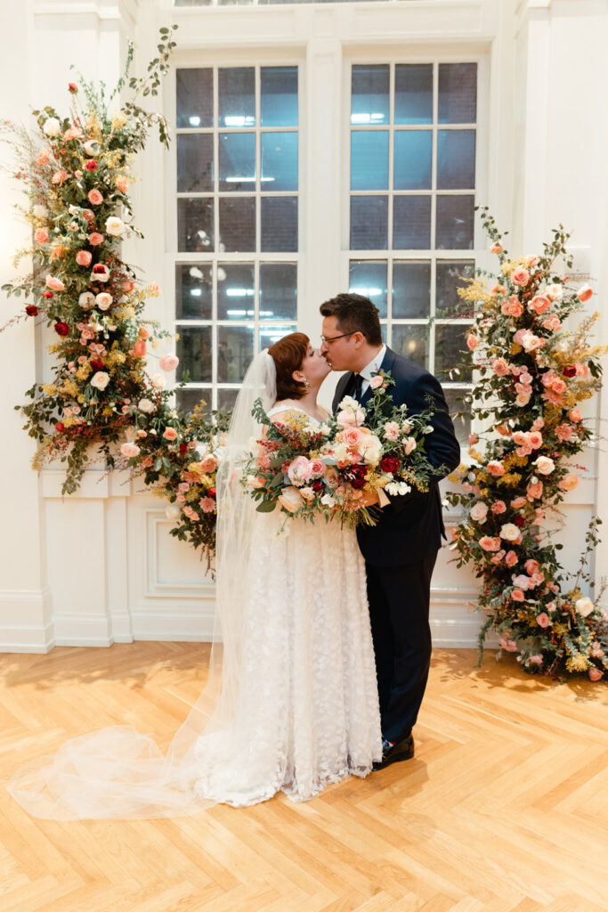 Asymmetrical growing floral installation for winter wedding in hues of mustard yellow, pink, burgundy, white, and sage green composed of petal heavy roses, acacia, mums, spray roses, butterfly ranunculus, ranunculus, and natural greenery. Design by Rosemary and Finch in Nashville, TN. 