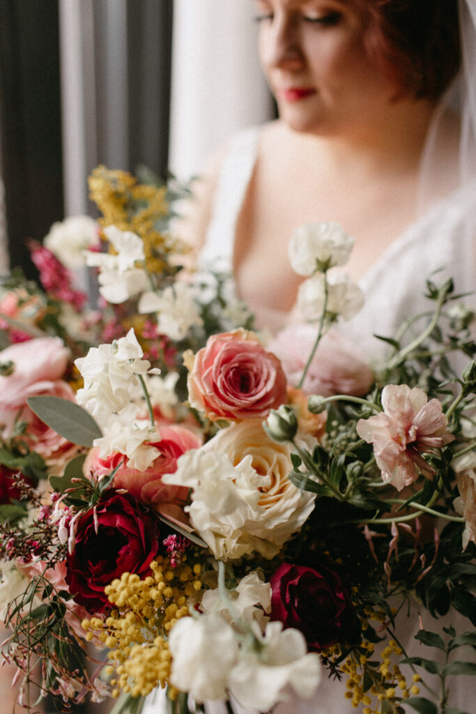 Lush winter bridal bouquet in floral hues of white, burgundy, mustard yellow, pink, and sage green composed of roses, sweet peas, spray roses, acacia, butterfly ranunculus, and eucalyptus. Design by Rosemary and Finch in Nashville, TN. 