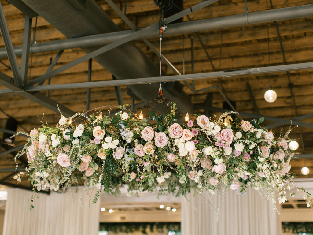 Spring wedding reception floral hanging chandelier in shades of cream, mauve, pink, lavender, and green featuring roses, spring floral branches, and greenery. Circular shaped hoop floral chandelier. Design by Rosemary and Finch Floral Design in Nashville, TN. 