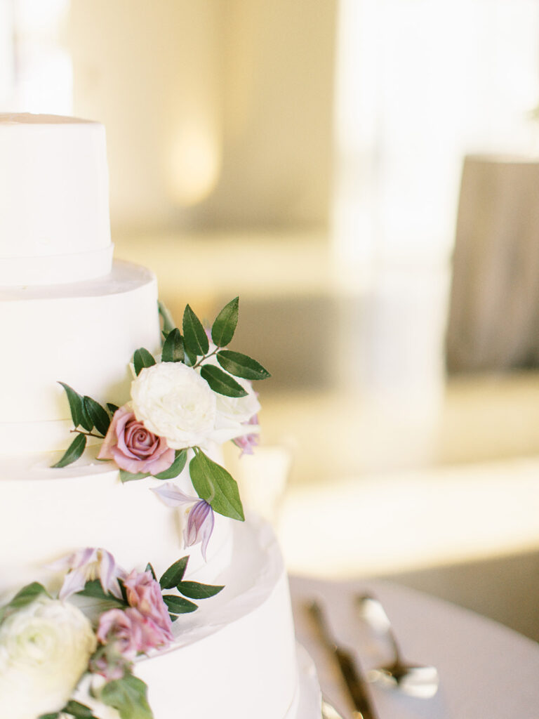 Elegant cake flowers add color to this all white classic wedding cake. Flower colors in pink, purple, lavender, white, cream, and green featuring roses, ranunculus, and greenery. Design by Rosemary and Finch Floral Design in Nashville, TN.