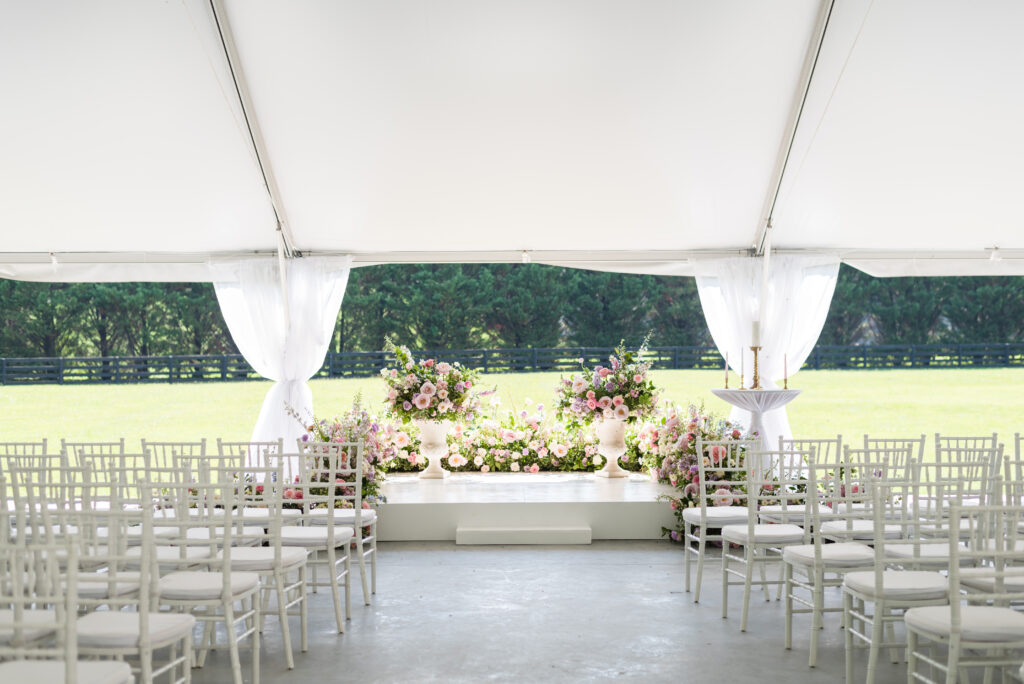Lush summer wedding ceremony backdrop floral meadows on platform stage with floral urns. Pastel flower colors in light pink, lavender, cream, blush, peach, and natural green consisting of large roses, ranunculus, and delphinium. Design by Rosemary and Finch at Cherokee Dock in Nashville, TN. 