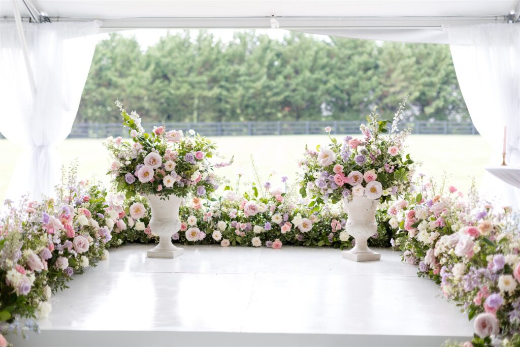 Lush summer wedding ceremony backdrop floral meadows on platform stage with floral urns. Pastel flower colors in light pink, lavender, cream, blush, peach, and natural green consisting of large roses, ranunculus, and delphinium. Design by Rosemary and Finch at Cherokee Dock in Nashville, TN. 