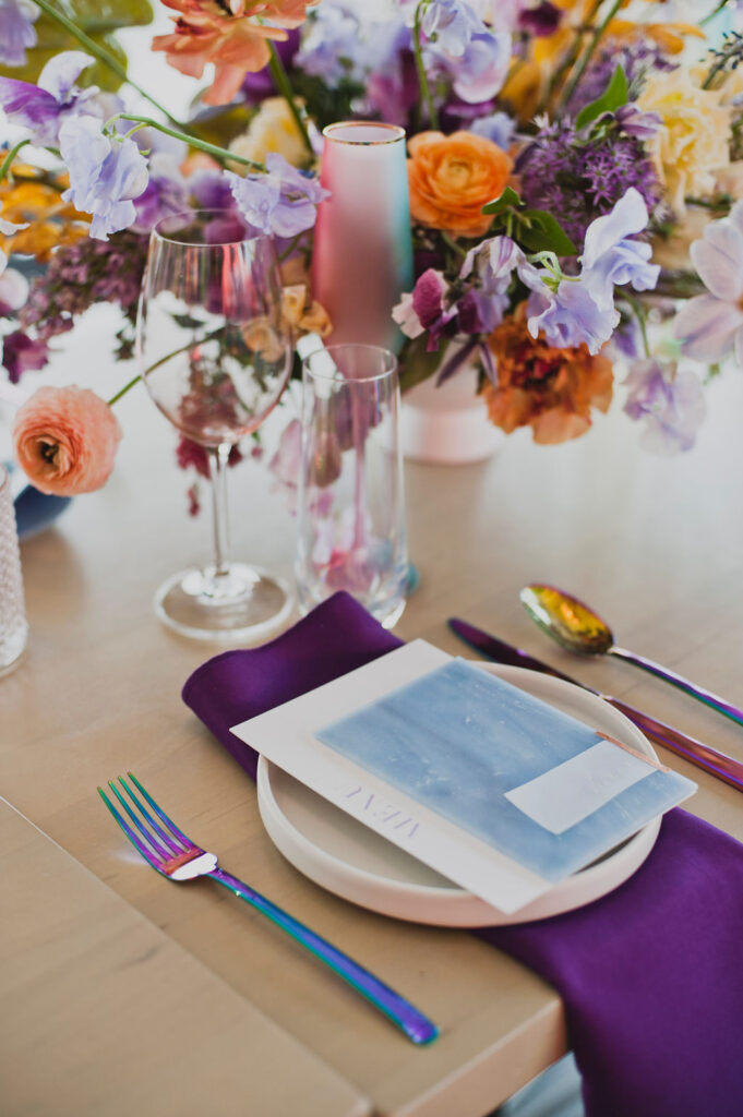 Iridescent colorful design is brought to life with these unique floral colors. Low floral centerpiece with colorful florals in purple, blue, orange, and yellow. Artful floral design in bold colors decorate this reception table for an exciting dinner party. Anemones, ranunculus, tulips, sweet peas, and anthurium bring a rainbow of color to this floral design. Paired with iridescent Estelle colored glassware, candles, place settings, cutlery, and florals. Bold floral design with artful shape and interesting color. Design by Rosemary & Finch Floral Design in Nashville, TN.