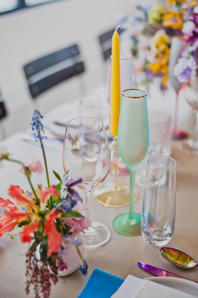 Colorful bud vases bring pop of color to this unique iridescent color palette reception. Unique and bright colors in pink, purple, blue, red, orange, and yellow. Tulips, sweet pea, and brightly colored wildflowers bring fun florals to brightly colored wedding reception. Paired with iridescent Estelle colored glassware, candles, place settings, cutlery, and florals. Bold floral design with artful shape and interesting color. Design by Rosemary & Finch in Nashville, TN.
