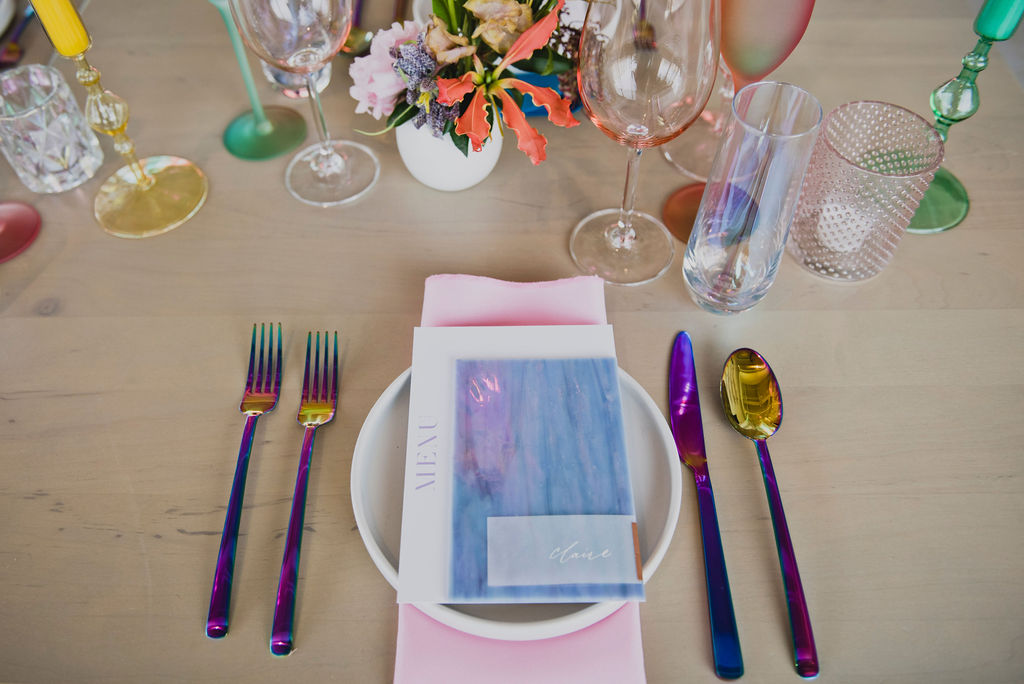 Colorful bud vases bring pop of color to this unique iridescent color palette reception. Unique and bright colors in pink, purple, blue, red, orange, and yellow. Tulips, sweet pea, and brightly colored wildflowers bring fun florals to brightly colored wedding reception. Paired with iridescent Estelle colored glassware, candles, place settings, cutlery, and florals. Bold floral design with artful shape and interesting color. Design by Rosemary & Finch in Nashville, TN.