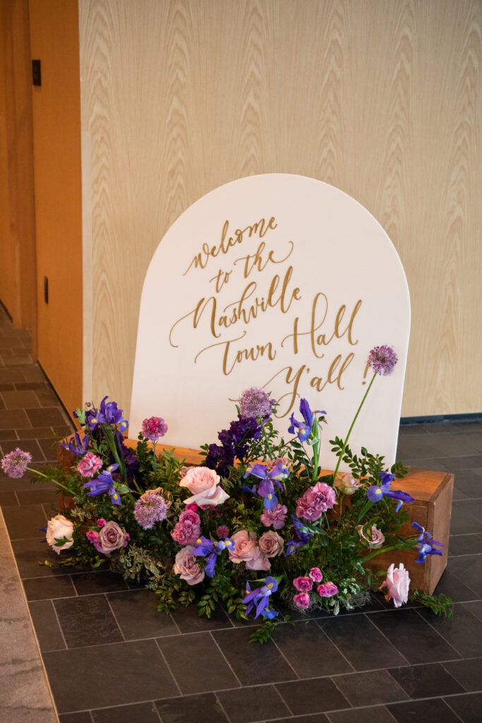 Welcome sign floral meadow brightens the entrance to this spring wedding industry event for WIPA Nashville. Floral colors of purple, pink, lavender, mauve, and natural greens frame the seating area with flowers. Corporate industry event florals in Downtown Nashville. Design by Rosemary and Finch Floral Design in Nashville, TN.