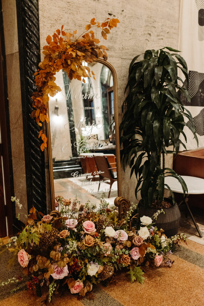 Tasteful fall wedding cocktail hour florals create an inviting space in hues of dusty rose, burgundy, mauve, taupe, copper, and lavender. Fall colors for bud vases, welcome sign, and bar of wedding cocktail hour. Florals composed of roses, clematis, lisianthus, spray roses, copper beech, and fall foliage. Fall wedding in Downtown Nashville. Design by Rosemary & Finch Floral. Design in Nashville, TN. 