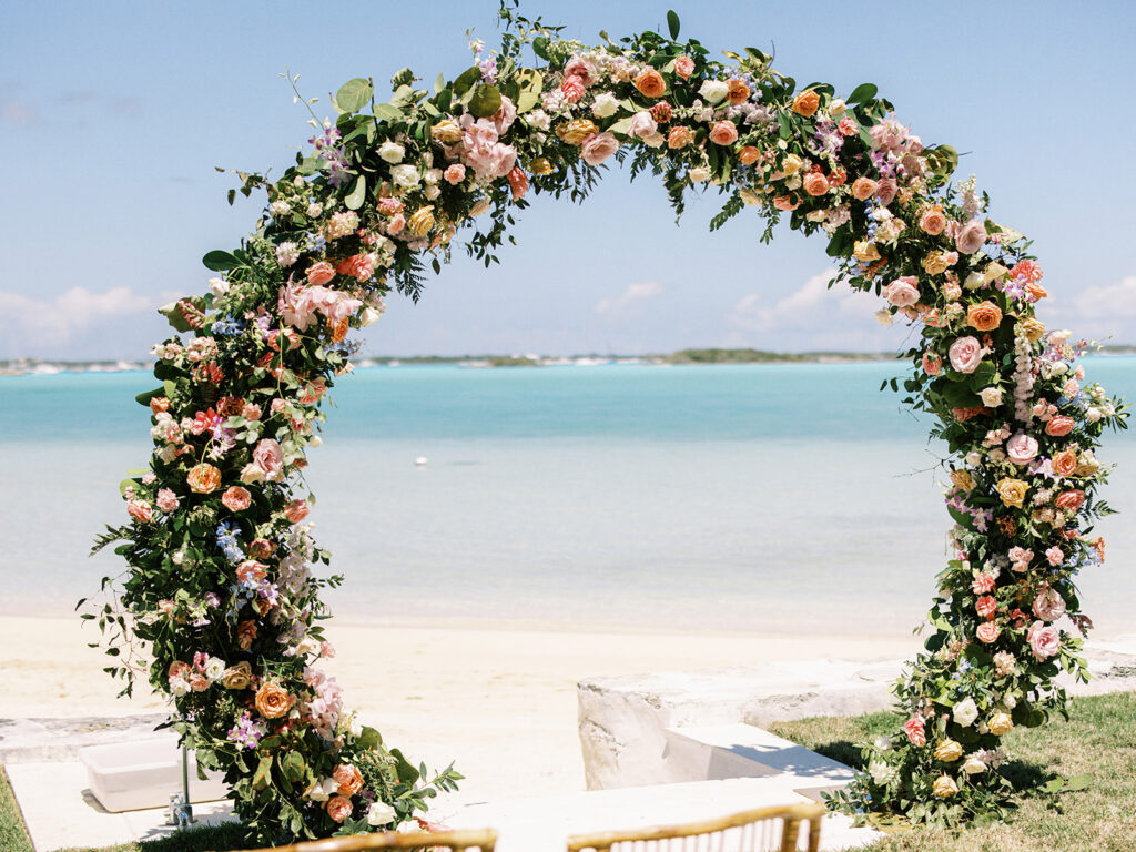 Double circle arch floral ceremony arch for spring tropical wedding ceremony. Exuma, Bahamas beach private estate destination beach wedding. Roses, tropical flowers, tropical foliage in colors of peach, dusty pink, orange, dusty blue, cream, lavender, and natural greenery. Floral Design by Rosemary & Finch Floral Design. 