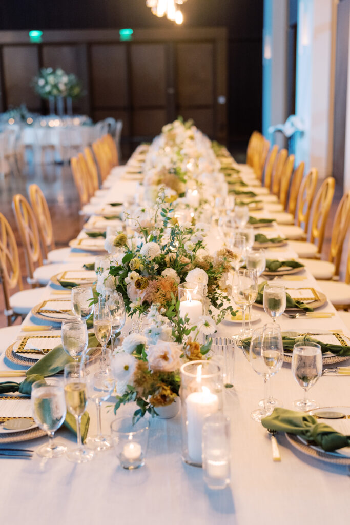 Lush collection of low centerpieces line this garden-inspired head table for summer wedding reception. Classic white and green wedding with floral colors in cream, white, taupe, and champagne. Summer floral design full of roses, ranunculus, hydrangea, cosmos, and lisianthis. Design by Rosemary & Finch Floral Design in Nashville, TN. 