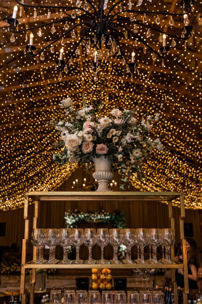 Glitzy wedding reception florals with twinkling lights for fall wedding. Statement urn adorns the reception bar with timeless floral colors in white, cream, and blush. Roses, anemones, and sweet peas decorate this fall destination wedding in Nashville, TN. Design by Rosemary & Finch Floral Design in Nashville, TN.