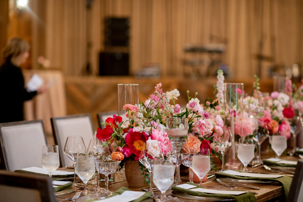A collection of floral heavy centerpieces fills the head table at this spring wedding with fun floral colors. Pink florals at vibrant color to this spring wedding reception with pink peonies, roses, ranunculus, and pink tulips. Lush florals and pink candles decorate this epic pink head table. Destination wedding in Nashville countryside. Design by Rosemary & Finch Floral Design in Nashville, TN.