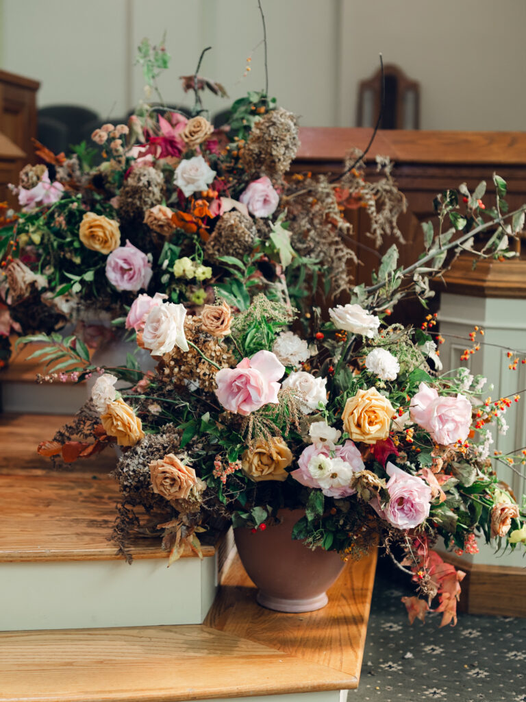 Large floral arrangements for ceremony backdrop. Staggered statement urn floral backdrop for fall wedding ceremony. Fall floral colors in dusty rose, copper, caramel, honey, and burgundy. Tasteful floral backdrop for church wedding ceremony with floral urns on either side. Destination wedding floral design for Nashville, TN. Design by Rosemary & Finch Floral Design in Nashville, TN. 