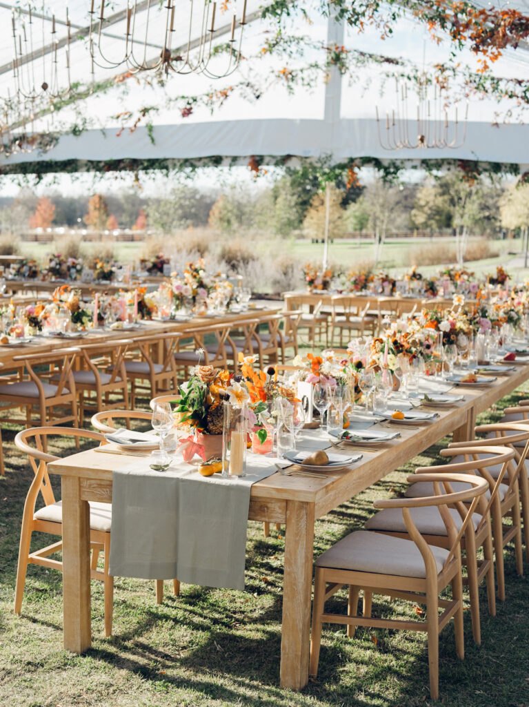 Full tablescapes of fall flowers create lush warm fall setting for wedding reception in Tennessee countryside. Wedding colors in blush, tangerine, mustard yellow, caramel, and honey. Wedding fall florals consisting of roses, clematis, ranunculus, brown tulips, and fall foliage. A collection of low centerpieces and accent arrangements create lush floral wedding reception tables with styled fruit place settings. Unique and colorful candles for reception tables in pink, orange, yellow, and gold. Destination floral design by Rosemary & Finch Floral Design in Nashville, TN.