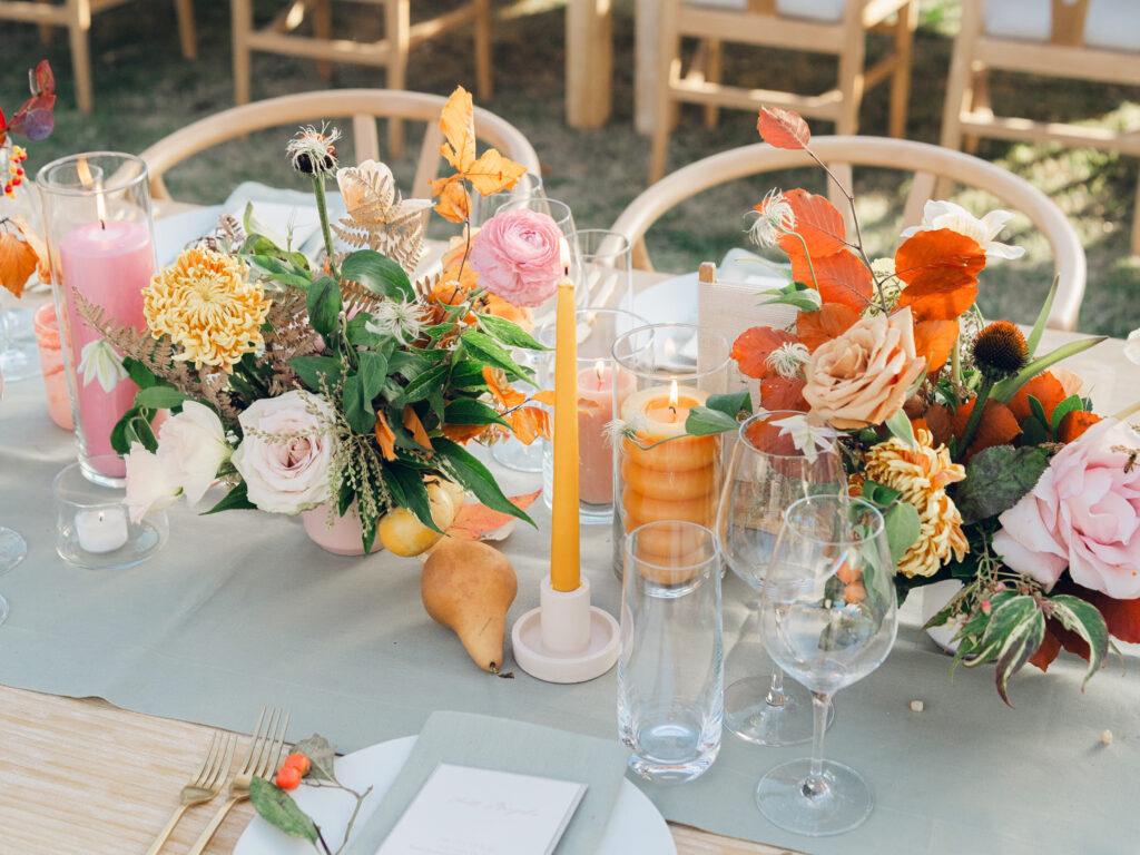 Full tablescapes of fall flowers create lush warm fall setting for wedding reception in Tennessee countryside. Wedding colors in blush, tangerine, mustard yellow, caramel, and honey. Wedding fall florals consisting of roses, clematis, ranunculus, brown tulips, and fall foliage. A collection of low centerpieces and accent arrangements create lush floral wedding reception tables with styled fruit place settings. Unique and colorful candles for reception tables in pink, orange, yellow, and gold. Destination floral design by Rosemary & Finch Floral Design in Nashville, TN.