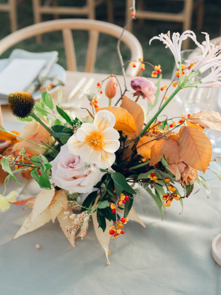 Full tablescapes of fall flowers create lush warm fall setting for wedding reception in Tennessee countryside. Wedding colors in blush, tangerine, mustard yellow, caramel, and honey. Wedding fall florals consisting of roses, clematis, ranunculus, brown tulips, and fall foliage. A collection of low centerpieces and accent arrangements create lush floral wedding reception tables with styled fruit place settings. Unique and colorful candles for reception tables in pink, orange, yellow, and gold. Destination floral design by Rosemary & Finch Floral Design in Nashville, TN.