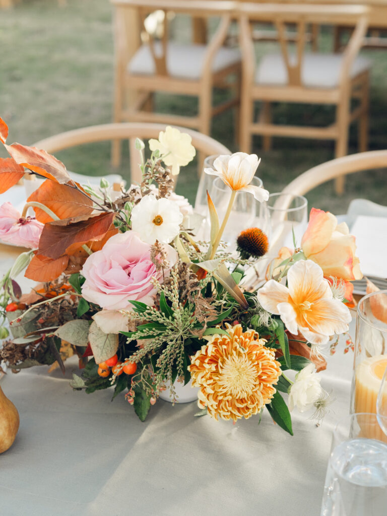 Full tablescapes of fall flowers create lush warm fall setting for wedding reception in Tennessee countryside. Wedding colors in blush, tangerine, mustard yellow, caramel, and honey. Wedding fall florals consisting of roses, clematis, ranunculus, brown tulips, and fall foliage. A collection of low centerpieces and accent arrangements create lush floral wedding reception tables with styled fruit place settings. Unique and colorful candles for reception tables in pink, orange, yellow, and gold. Destination floral design by Rosemary & Finch Floral Design in Nashville, TN.