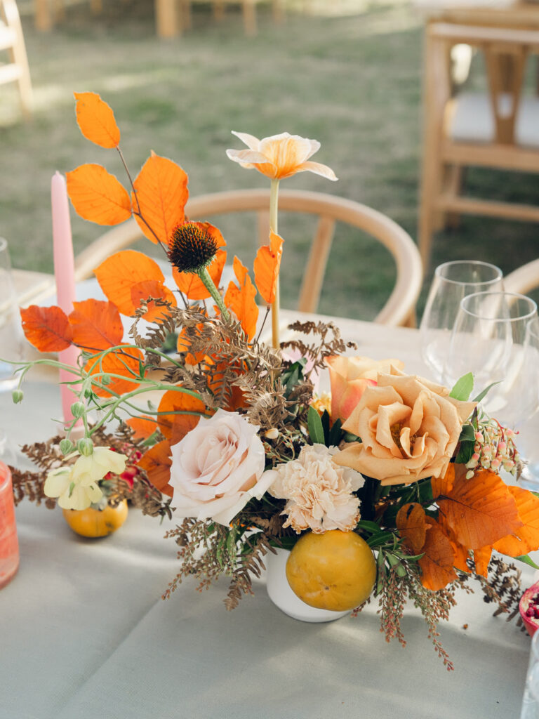 Full tablescapes of fall flowers create lush warm fall setting for wedding reception in Tennessee countryside. Wedding colors in blush, tangerine, mustard yellow, caramel, and honey. Wedding fall florals consisting of roses, clematis, ranunculus, brown tulips, and fall foliage. A collection of low centerpieces and accent arrangements create lush floral wedding reception tables with styled fruit place settings. Unique and colorful candles for reception tables in pink, orange, yellow, and gold. Destination floral design by Rosemary & Finch Floral Design in Nashville, TN.