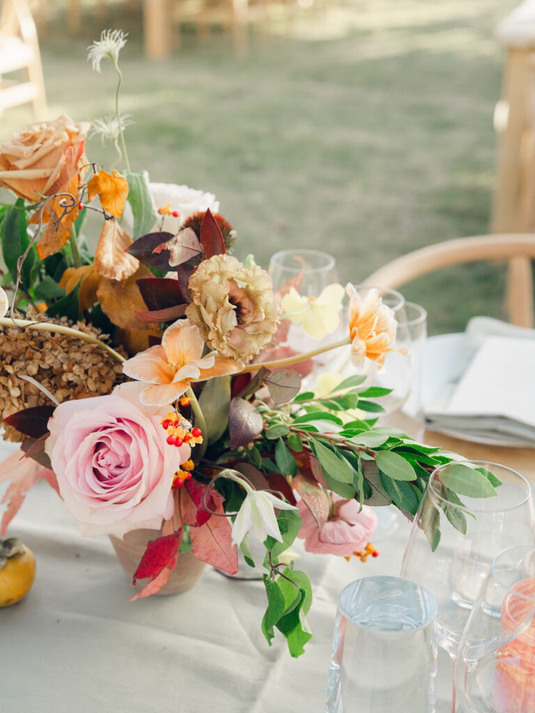 Full tablescapes of fall flowers create lush warm fall setting for wedding reception in Tennessee countryside. Wedding colors in blush, tangerine, mustard yellow, caramel, and honey. Wedding fall florals consisting of roses, clematis, ranunculus, brown tulips, and fall foliage. A collection of low centerpieces and accent arrangements create lush floral wedding reception tables with styled fruit place settings. Unique and colorful candles for reception tables in pink, orange, yellow, and gold. Destination floral design by Rosemary & Finch Floral Design in Nashville, TN.