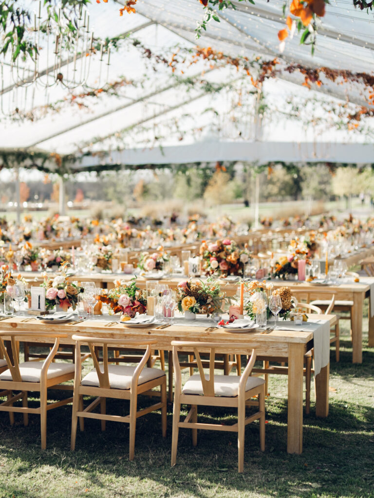 Full tablescapes of fall flowers create lush warm fall setting for wedding reception in Tennessee countryside. Wedding colors in blush, tangerine, mustard yellow, caramel, and honey. Wedding fall florals consisting of roses, clematis, ranunculus, brown tulips, and fall foliage. A collection of low centerpieces and accent arrangements create lush floral wedding reception tables with styled fruit place settings. Unique and colorful candles for reception tables in pink, orange, yellow, and gold. Destination floral design by Rosemary & Finch Floral Design in Nashville, TN.