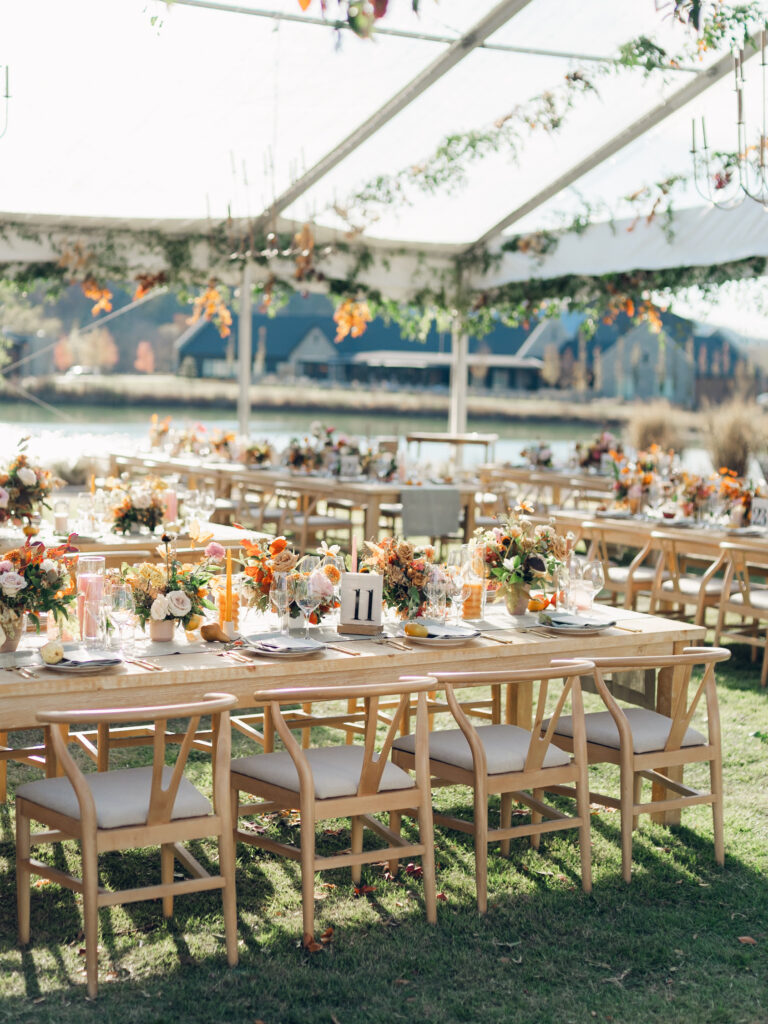 Full tablescapes of fall flowers create lush warm fall setting for wedding reception in Tennessee countryside. Wedding colors in blush, tangerine, mustard yellow, caramel, and honey. Wedding fall florals consisting of roses, clematis, ranunculus, brown tulips, and fall foliage. A collection of low centerpieces and accent arrangements create lush floral wedding reception tables with styled fruit place settings. Unique and colorful candles for reception tables in pink, orange, yellow, and gold. Destination floral design by Rosemary & Finch Floral Design in Nashville, TN.