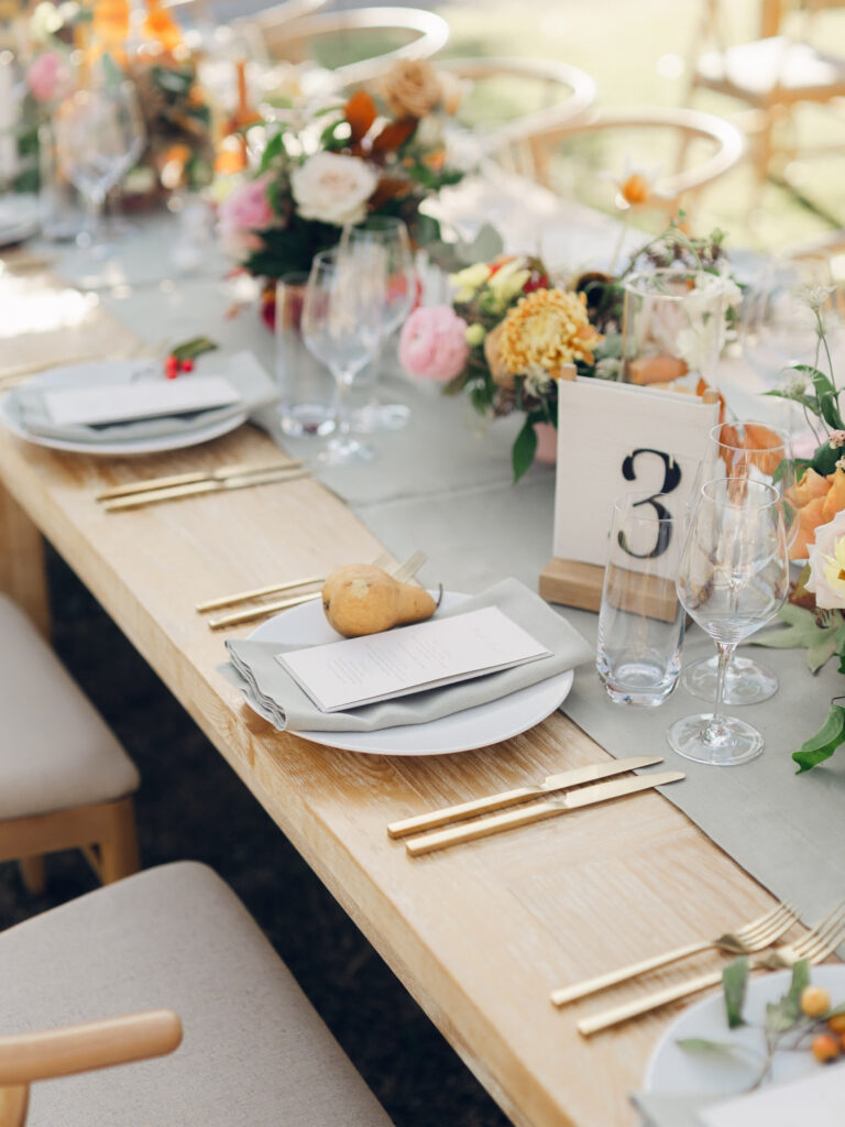 Full tablescapes of fall flowers create lush warm fall setting for wedding reception in Tennessee countryside. Wedding colors in blush, tangerine, mustard yellow, caramel, and honey. Wedding fall florals consisting of roses, clematis, ranunculus, brown tulips, and fall foliage. A collection of low centerpieces and accent arrangements create lush floral wedding reception tables with styled fruit place settings. Unique and colorful candles for reception tables in pink, orange, yellow, and gold. Destination floral design by Rosemary & Finch Floral Design in Nashville, TN.