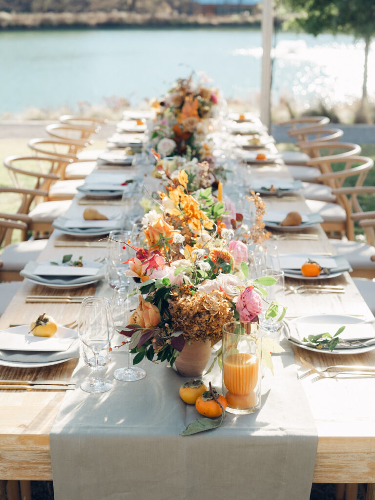 Full tablescapes of fall flowers create lush warm fall setting for wedding reception in Tennessee countryside. Wedding colors in blush, tangerine, mustard yellow, caramel, and honey. Wedding fall florals consisting of roses, clematis, ranunculus, brown tulips, and fall foliage. A collection of low centerpieces and accent arrangements create lush floral wedding reception tables with styled fruit place settings. Unique and colorful candles for reception tables in pink, orange, yellow, and gold. Destination floral design by Rosemary & Finch Floral Design in Nashville, TN.