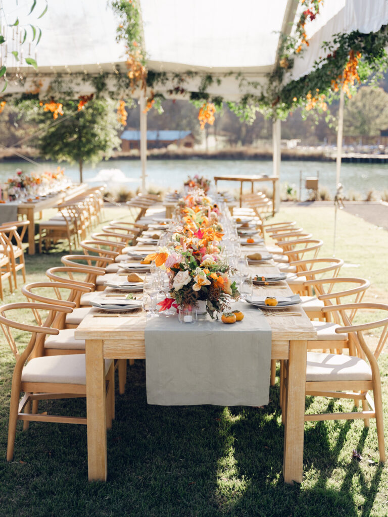 Full tablescapes of fall flowers create lush warm fall setting for wedding reception in Tennessee countryside. Wedding colors in blush, tangerine, mustard yellow, caramel, and honey. Wedding fall florals consisting of roses, clematis, ranunculus, brown tulips, and fall foliage. A collection of low centerpieces and accent arrangements create lush floral wedding reception tables with styled fruit place settings. Unique and colorful candles for reception tables in pink, orange, yellow, and gold. Destination floral design by Rosemary & Finch Floral Design in Nashville, TN.