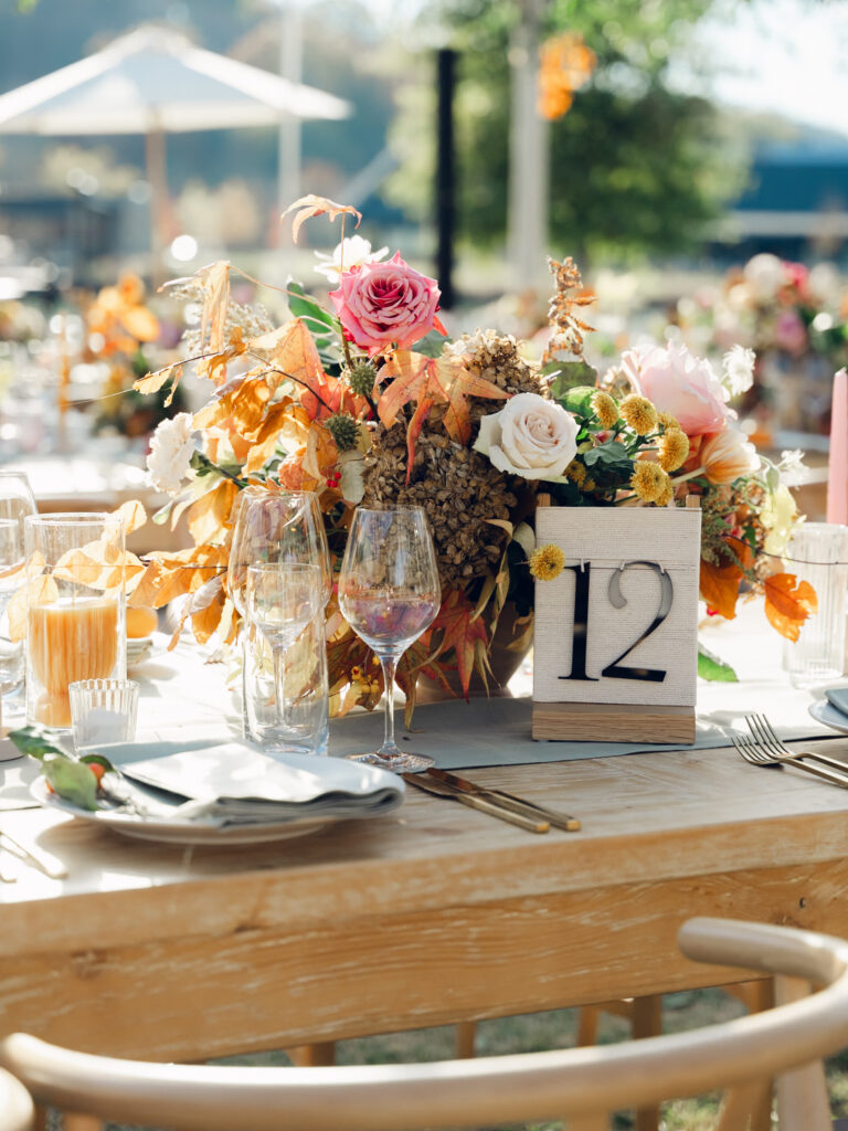 Full tablescapes of fall flowers create lush warm fall setting for wedding reception in Tennessee countryside. Wedding colors in blush, tangerine, mustard yellow, caramel, and honey. Wedding fall florals consisting of roses, clematis, ranunculus, brown tulips, and fall foliage. A collection of low centerpieces and accent arrangements create lush floral wedding reception tables with styled fruit place settings. Unique and colorful candles for reception tables in pink, orange, yellow, and gold. Destination floral design by Rosemary & Finch Floral Design in Nashville, TN.