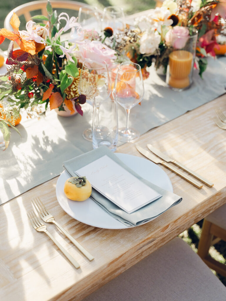 Full tablescapes of fall flowers create lush warm fall setting for wedding reception in Tennessee countryside. Wedding colors in blush, tangerine, mustard yellow, caramel, and honey. Wedding fall florals consisting of roses, clematis, ranunculus, brown tulips, and fall foliage. A collection of low centerpieces and accent arrangements create lush floral wedding reception tables with styled fruit place settings. Unique and colorful candles for reception tables in pink, orange, yellow, and gold. Destination floral design by Rosemary & Finch Floral Design in Nashville, TN.