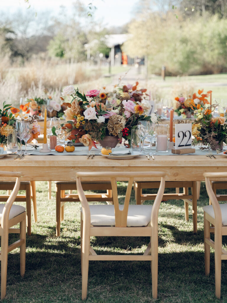 Full tablescapes of fall flowers create lush warm fall setting for wedding reception in Tennessee countryside. Wedding colors in blush, tangerine, mustard yellow, caramel, and honey. Wedding fall florals consisting of roses, clematis, ranunculus, brown tulips, and fall foliage. A collection of low centerpieces and accent arrangements create lush floral wedding reception tables with styled fruit place settings. Unique and colorful candles for reception tables in pink, orange, yellow, and gold. Destination floral design by Rosemary & Finch Floral Design in Nashville, TN.