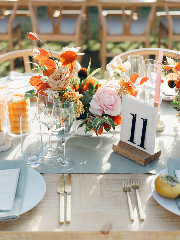 Full tablescapes of fall flowers create lush warm fall setting for wedding reception in Tennessee countryside. Wedding colors in blush, tangerine, mustard yellow, caramel, and honey. Wedding fall florals consisting of roses, clematis, ranunculus, brown tulips, and fall foliage. A collection of low centerpieces and accent arrangements create lush floral wedding reception tables with styled fruit place settings. Unique and colorful candles for reception tables in pink, orange, yellow, and gold. Destination floral design by Rosemary & Finch Floral Design in Nashville, TN.