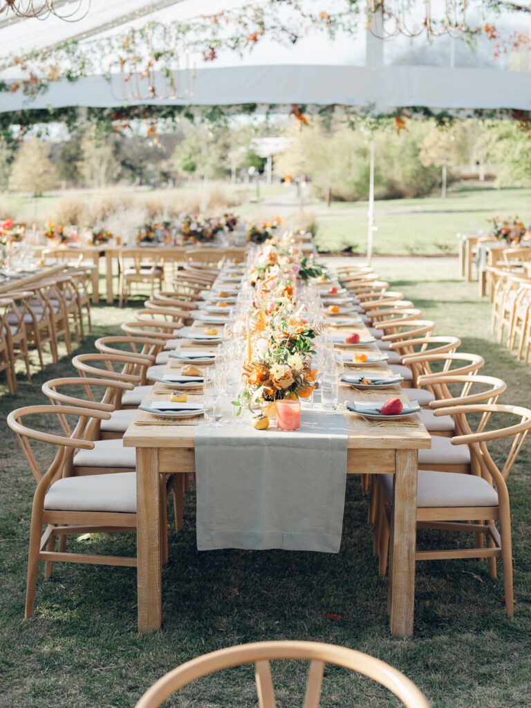 Full tablescapes of fall flowers create lush warm fall setting for wedding reception in Tennessee countryside. Wedding colors in blush, tangerine, mustard yellow, caramel, and honey. Wedding fall florals consisting of roses, clematis, ranunculus, brown tulips, and fall foliage. A collection of low centerpieces and accent arrangements create lush floral wedding reception tables with styled fruit place settings. Unique and colorful candles for reception tables in pink, orange, yellow, and gold. Destination floral design by Rosemary & Finch Floral Design in Nashville, TN.