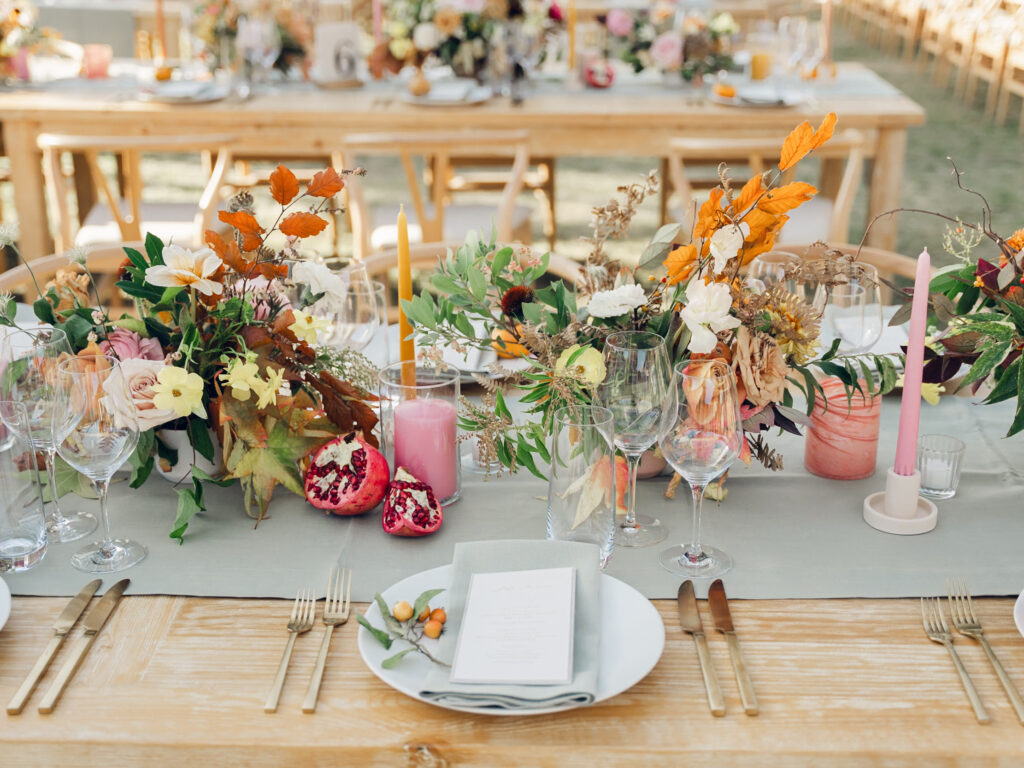 Full tablescapes of fall flowers create lush warm fall setting for wedding reception in Tennessee countryside. Wedding colors in blush, tangerine, mustard yellow, caramel, and honey. Wedding fall florals consisting of roses, clematis, ranunculus, brown tulips, and fall foliage. A collection of low centerpieces and accent arrangements create lush floral wedding reception tables with styled fruit place settings. Unique and colorful candles for reception tables in pink, orange, yellow, and gold. Destination floral design by Rosemary & Finch Floral Design in Nashville, TN.