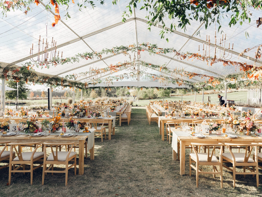 Full tablescapes of fall flowers create lush warm fall setting for wedding reception in Tennessee countryside. Wedding colors in blush, tangerine, mustard yellow, caramel, and honey. Wedding fall florals consisting of roses, clematis, ranunculus, brown tulips, and fall foliage. A collection of low centerpieces and accent arrangements create lush floral wedding reception tables with styled fruit place settings. Unique and colorful candles for reception tables in pink, orange, yellow, and gold. Destination floral design by Rosemary & Finch Floral Design in Nashville, TN.