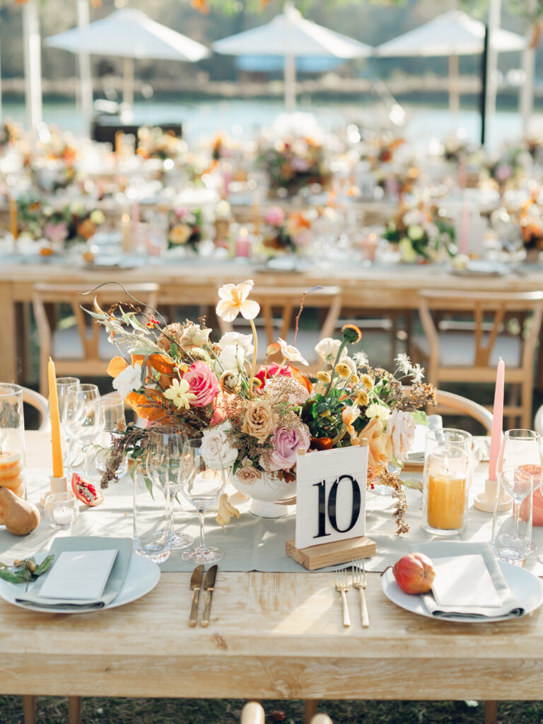 Full tablescapes of fall flowers create lush warm fall setting for wedding reception in Tennessee countryside. Wedding colors in blush, tangerine, mustard yellow, caramel, and honey. Wedding fall florals consisting of roses, clematis, ranunculus, brown tulips, and fall foliage. A collection of low centerpieces and accent arrangements create lush floral wedding reception tables with styled fruit place settings. Unique and colorful candles for reception tables in pink, orange, yellow, and gold. Destination floral design by Rosemary & Finch Floral Design in Nashville, TN.