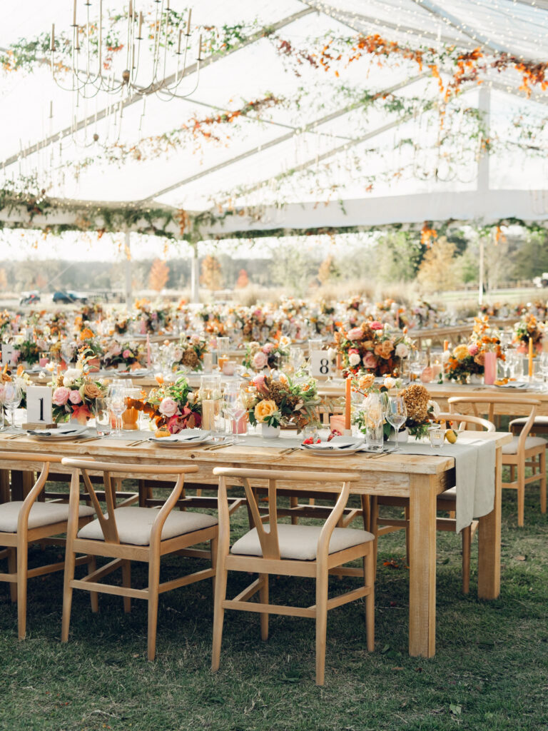 Full tablescapes of fall flowers create lush warm fall setting for wedding reception in Tennessee countryside. Wedding colors in blush, tangerine, mustard yellow, caramel, and honey. Wedding fall florals consisting of roses, clematis, ranunculus, brown tulips, and fall foliage. A collection of low centerpieces and accent arrangements create lush floral wedding reception tables with styled fruit place settings. Unique and colorful candles for reception tables in pink, orange, yellow, and gold. Destination floral design by Rosemary & Finch Floral Design in Nashville, TN.