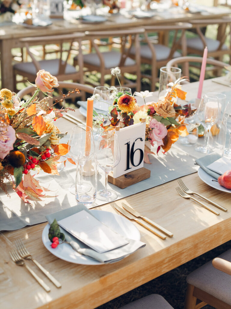 Full tablescapes of fall flowers create lush warm fall setting for wedding reception in Tennessee countryside. Wedding colors in blush, tangerine, mustard yellow, caramel, and honey. Wedding fall florals consisting of roses, clematis, ranunculus, brown tulips, and fall foliage. A collection of low centerpieces and accent arrangements create lush floral wedding reception tables with styled fruit place settings. Unique and colorful candles for reception tables in pink, orange, yellow, and gold. Destination floral design by Rosemary & Finch Floral Design in Nashville, TN.