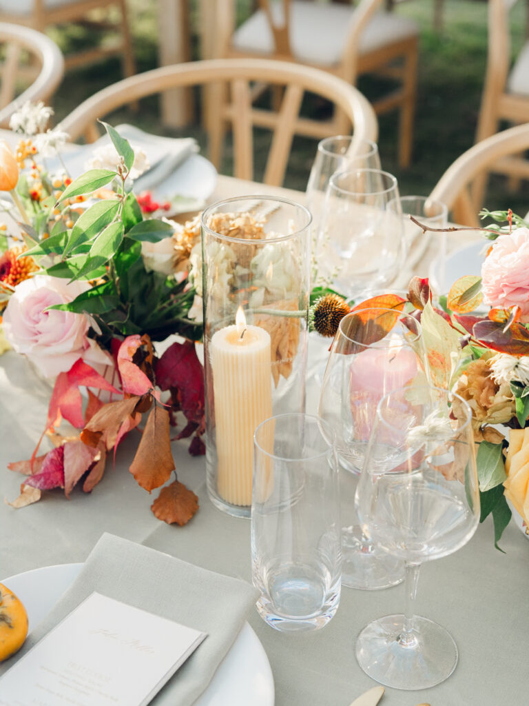 Full tablescapes of fall flowers create lush warm fall setting for wedding reception in Tennessee countryside. Wedding colors in blush, tangerine, mustard yellow, caramel, and honey. Wedding fall florals consisting of roses, clematis, ranunculus, brown tulips, and fall foliage. A collection of low centerpieces and accent arrangements create lush floral wedding reception tables with styled fruit place settings. Unique and colorful candles for reception tables in pink, orange, yellow, and gold. Destination floral design by Rosemary & Finch Floral Design in Nashville, TN.