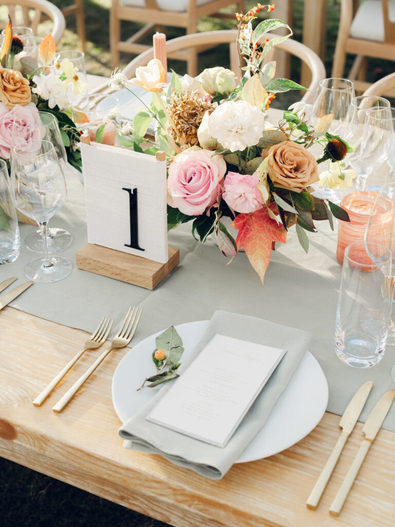 Full tablescapes of fall flowers create lush warm fall setting for wedding reception in Tennessee countryside. Wedding colors in blush, tangerine, mustard yellow, caramel, and honey. Wedding fall florals consisting of roses, clematis, ranunculus, brown tulips, and fall foliage. A collection of low centerpieces and accent arrangements create lush floral wedding reception tables with styled fruit place settings. Unique and colorful candles for reception tables in pink, orange, yellow, and gold. Destination floral design by Rosemary & Finch Floral Design in Nashville, TN.