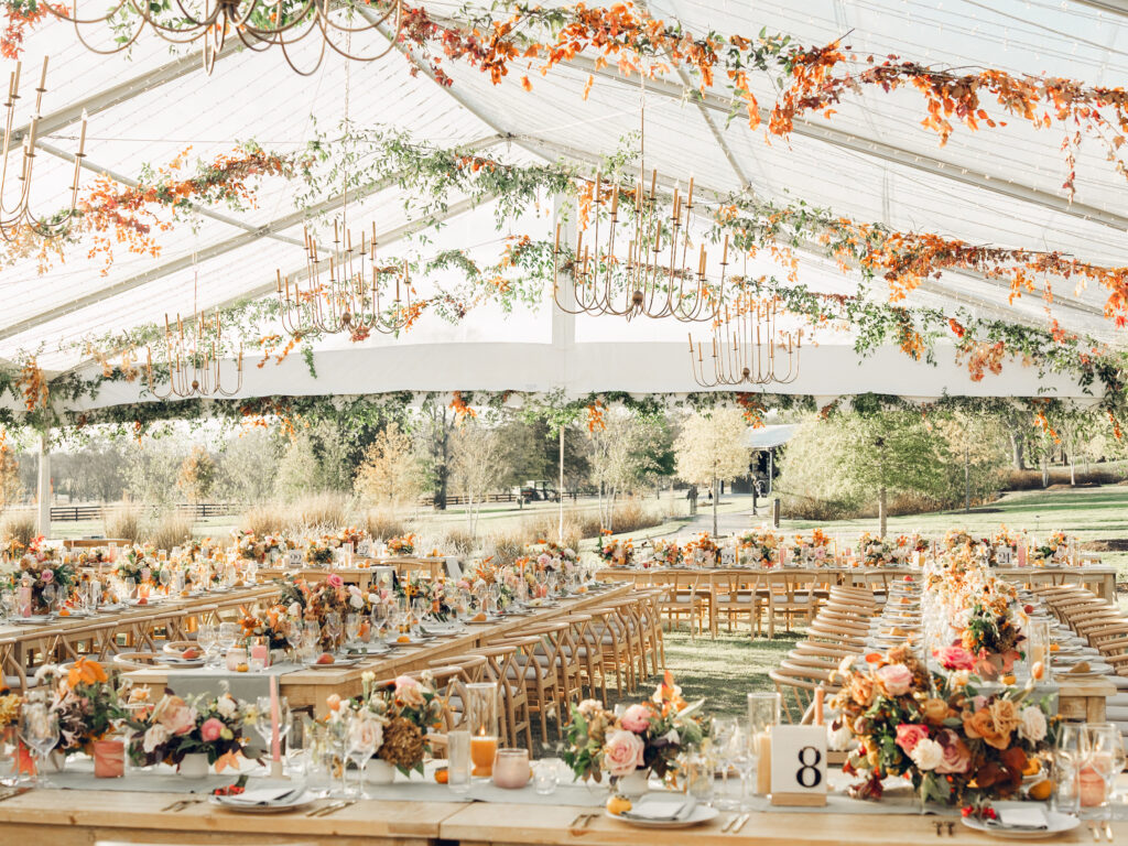 Full tablescapes of fall flowers create lush warm fall setting for wedding reception in Tennessee countryside. Wedding colors in blush, tangerine, mustard yellow, caramel, and honey. Wedding fall florals consisting of roses, clematis, ranunculus, brown tulips, and fall foliage. A collection of low centerpieces and accent arrangements create lush floral wedding reception tables with styled fruit place settings. Unique and colorful candles for reception tables in pink, orange, yellow, and gold. Destination floral design by Rosemary & Finch Floral Design in Nashville, TN.