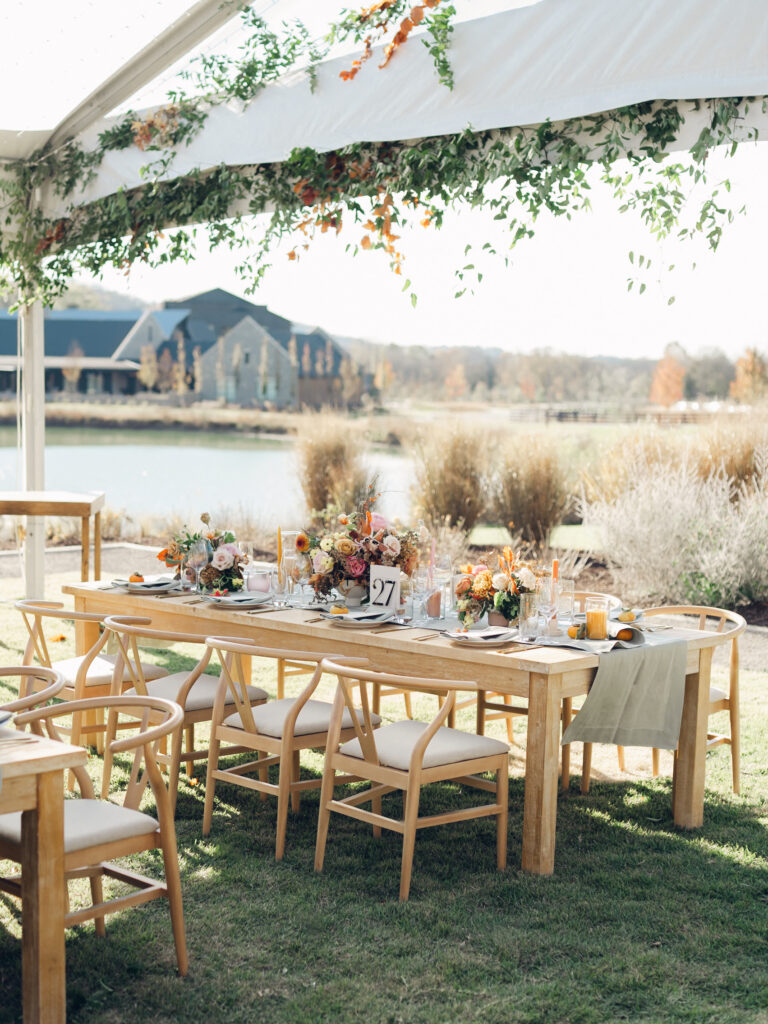 Full tablescapes of fall flowers create lush warm fall setting for wedding reception in Tennessee countryside. Wedding colors in blush, tangerine, mustard yellow, caramel, and honey. Wedding fall florals consisting of roses, clematis, ranunculus, brown tulips, and fall foliage. A collection of low centerpieces and accent arrangements create lush floral wedding reception tables with styled fruit place settings. Unique and colorful candles for reception tables in pink, orange, yellow, and gold. Destination floral design by Rosemary & Finch Floral Design in Nashville, TN.