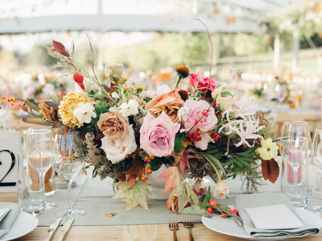 Full tablescapes of fall flowers create lush warm fall setting for wedding reception in Tennessee countryside. Wedding colors in blush, tangerine, mustard yellow, caramel, and honey. Wedding fall florals consisting of roses, clematis, ranunculus, brown tulips, and fall foliage. A collection of low centerpieces and accent arrangements create lush floral wedding reception tables with styled fruit place settings. Unique and colorful candles for reception tables in pink, orange, yellow, and gold. Destination floral design by Rosemary & Finch Floral Design in Nashville, TN.