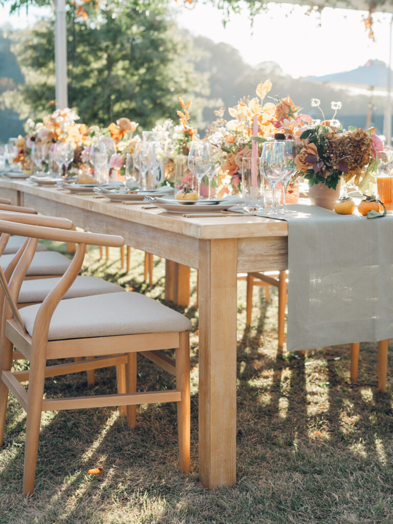 Full tablescapes of fall flowers create lush warm fall setting for wedding reception in Tennessee countryside. Wedding colors in blush, tangerine, mustard yellow, caramel, and honey. Wedding fall florals consisting of roses, clematis, ranunculus, brown tulips, and fall foliage. A collection of low centerpieces and accent arrangements create lush floral wedding reception tables with styled fruit place settings. Unique and colorful candles for reception tables in pink, orange, yellow, and gold. Destination floral design by Rosemary & Finch Floral Design in Nashville, TN.