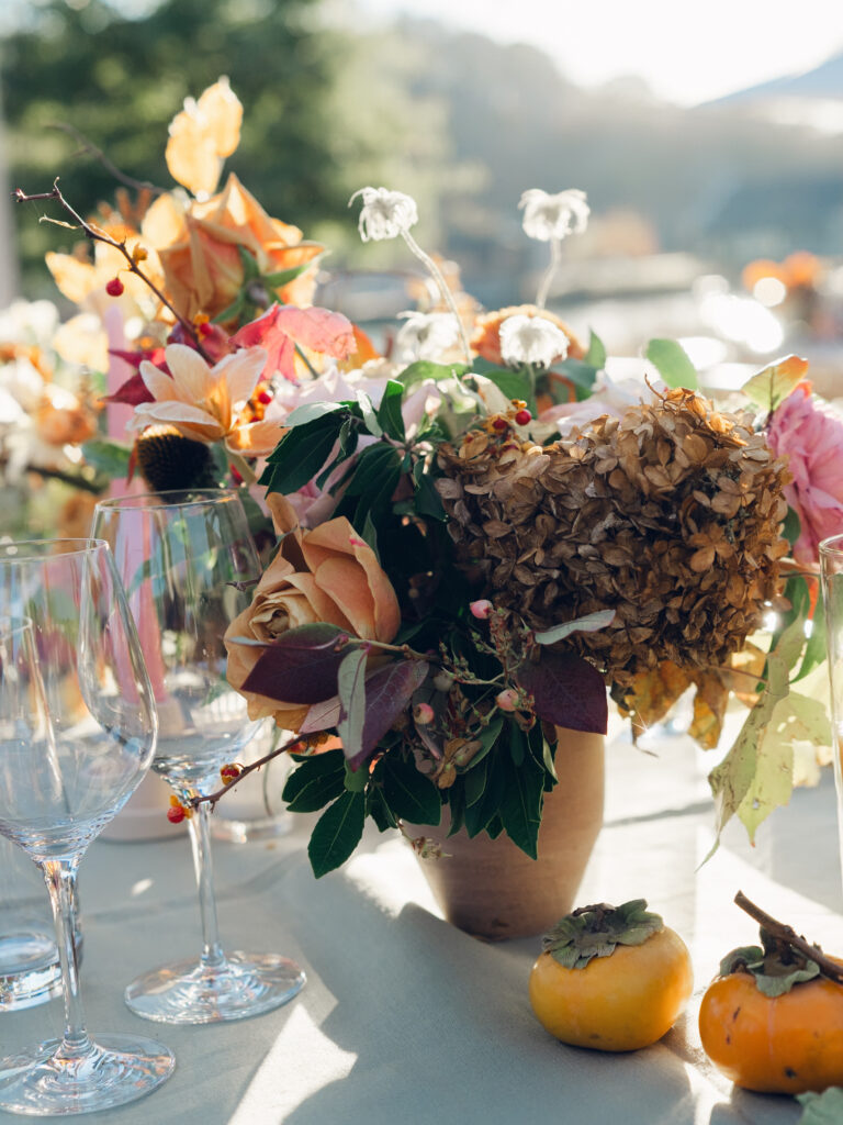 Full tablescapes of fall flowers create lush warm fall setting for wedding reception in Tennessee countryside. Wedding colors in blush, tangerine, mustard yellow, caramel, and honey. Wedding fall florals consisting of roses, clematis, ranunculus, brown tulips, and fall foliage. A collection of low centerpieces and accent arrangements create lush floral wedding reception tables with styled fruit place settings. Unique and colorful candles for reception tables in pink, orange, yellow, and gold. Destination floral design by Rosemary & Finch Floral Design in Nashville, TN.