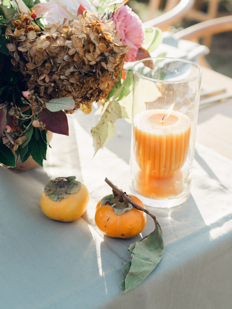 Full tablescapes of fall flowers create lush warm fall setting for wedding reception in Tennessee countryside. Wedding colors in blush, tangerine, mustard yellow, caramel, and honey. Wedding fall florals consisting of roses, clematis, ranunculus, brown tulips, and fall foliage. A collection of low centerpieces and accent arrangements create lush floral wedding reception tables with styled fruit place settings. Unique and colorful candles for reception tables in pink, orange, yellow, and gold. Destination floral design by Rosemary & Finch Floral Design in Nashville, TN.